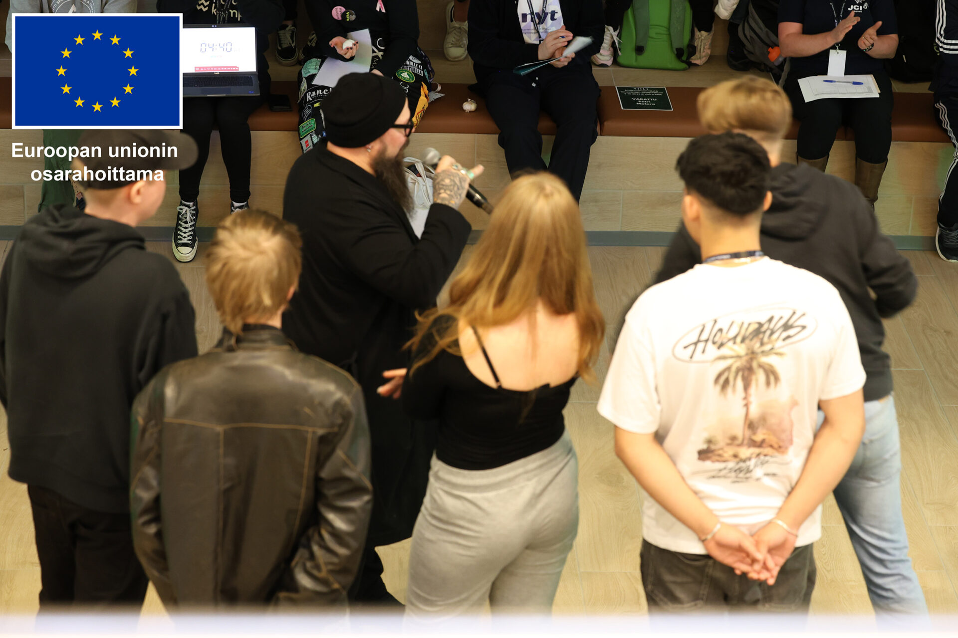 Neljä opiskelijaa ja juontaja lavalla, selkä kameraan päin. Taustalla näkyy yleisöä. Kuvan yläkulmassa Euroopan unionin osarahoittama -logo