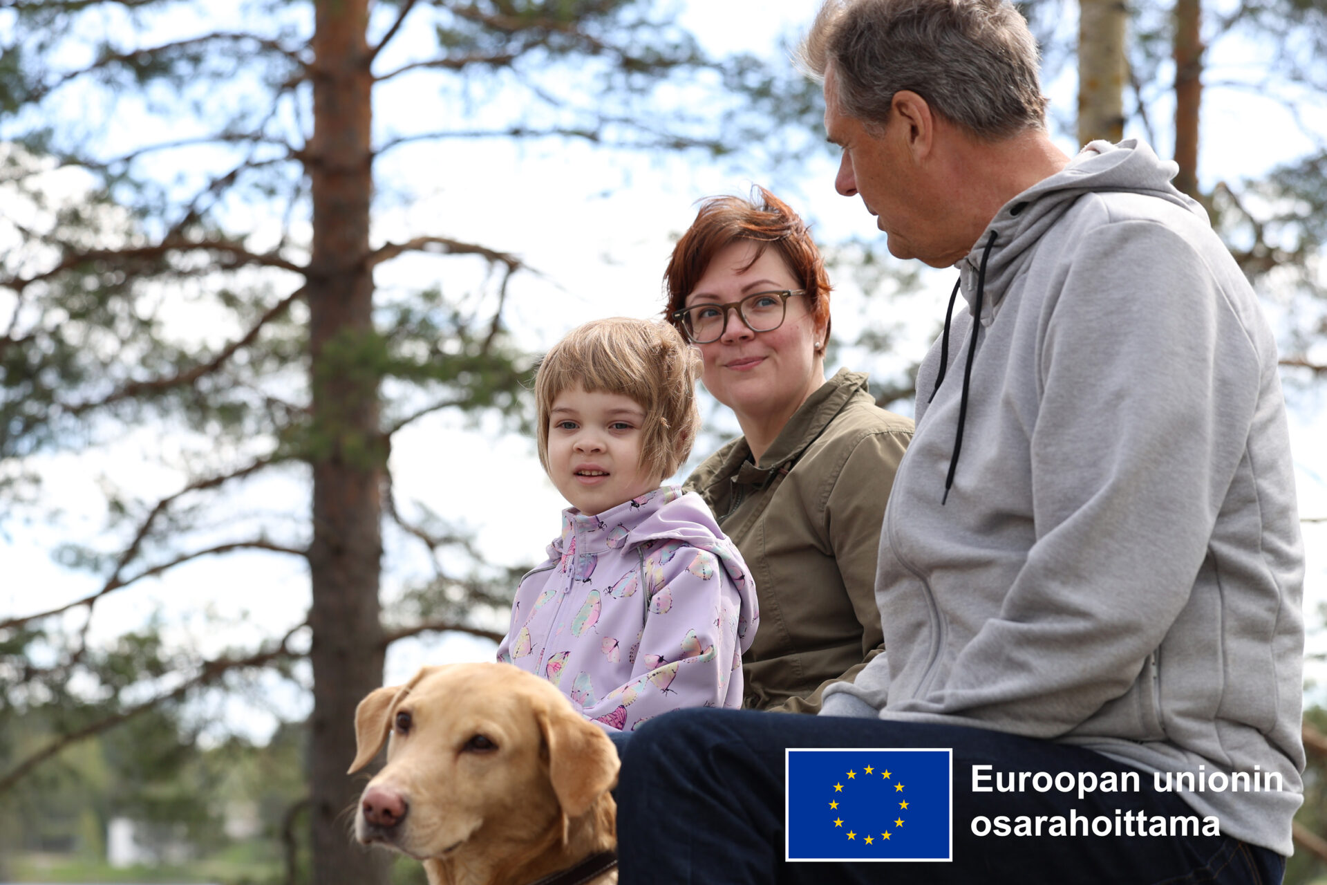 Vanha mies, keski-ikäinen nainen, lapsi ja koira istumassa metsässä. Alakulmassa Euroopan unionin osarahoittama -logo
