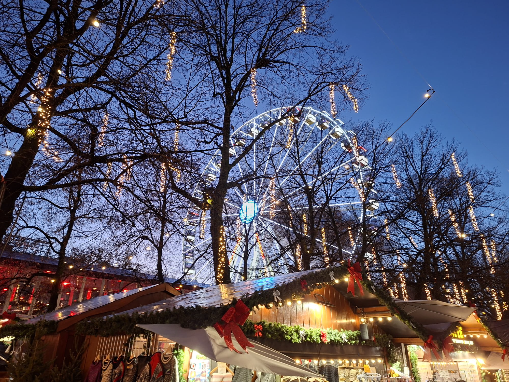 Trees above the christmas market in Karl Johans Gate, Oslo city center. The trees have lights hanging from them. Below the roof of a booth can be seen.