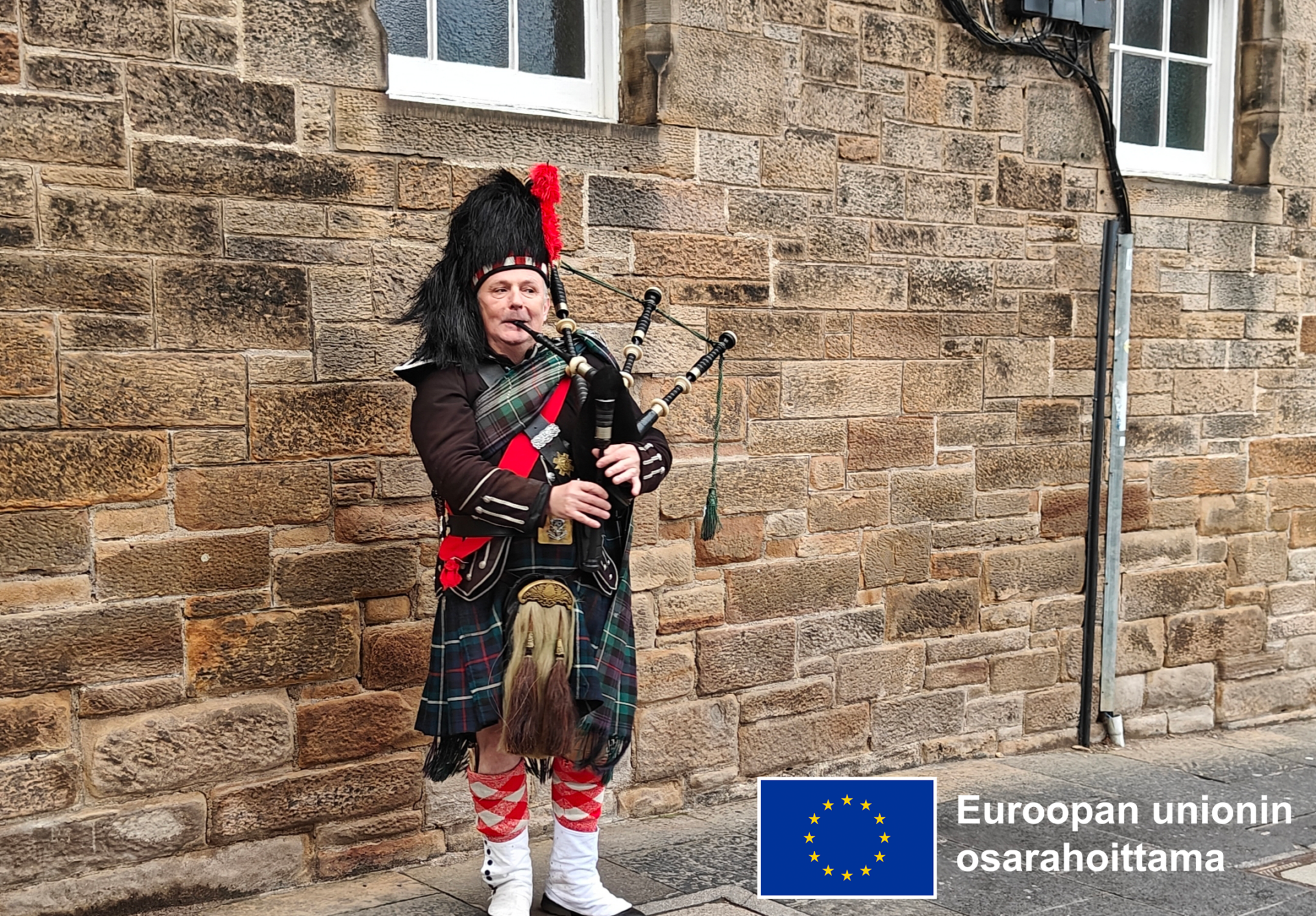 Säkkipillinsoittaja juhla-asussa kadulla. Kuvan alakulmassa Euroopan unionin osarahoittama -logo