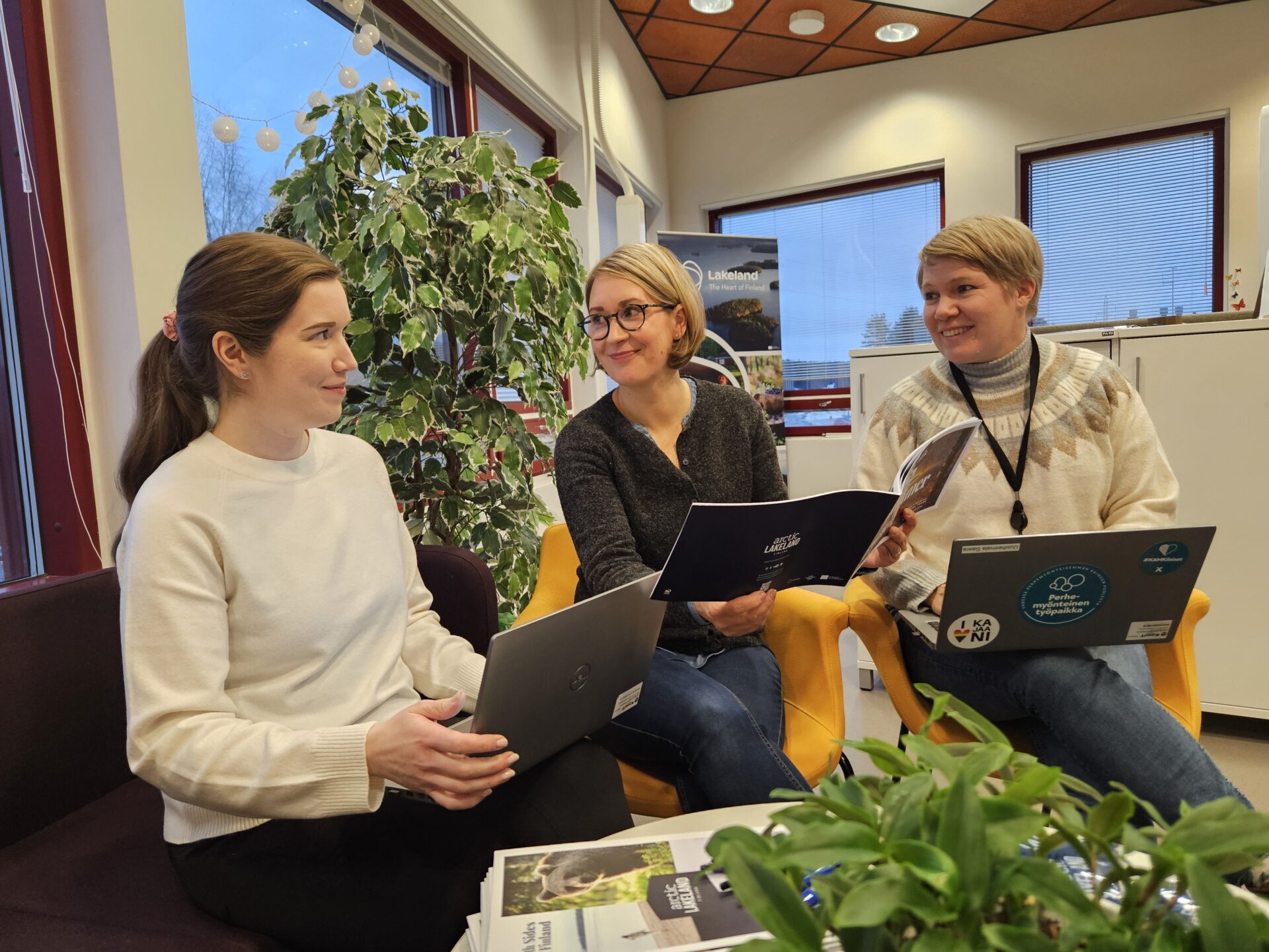 3 women sitting on a couch, holding laptops and a brochure in an office space