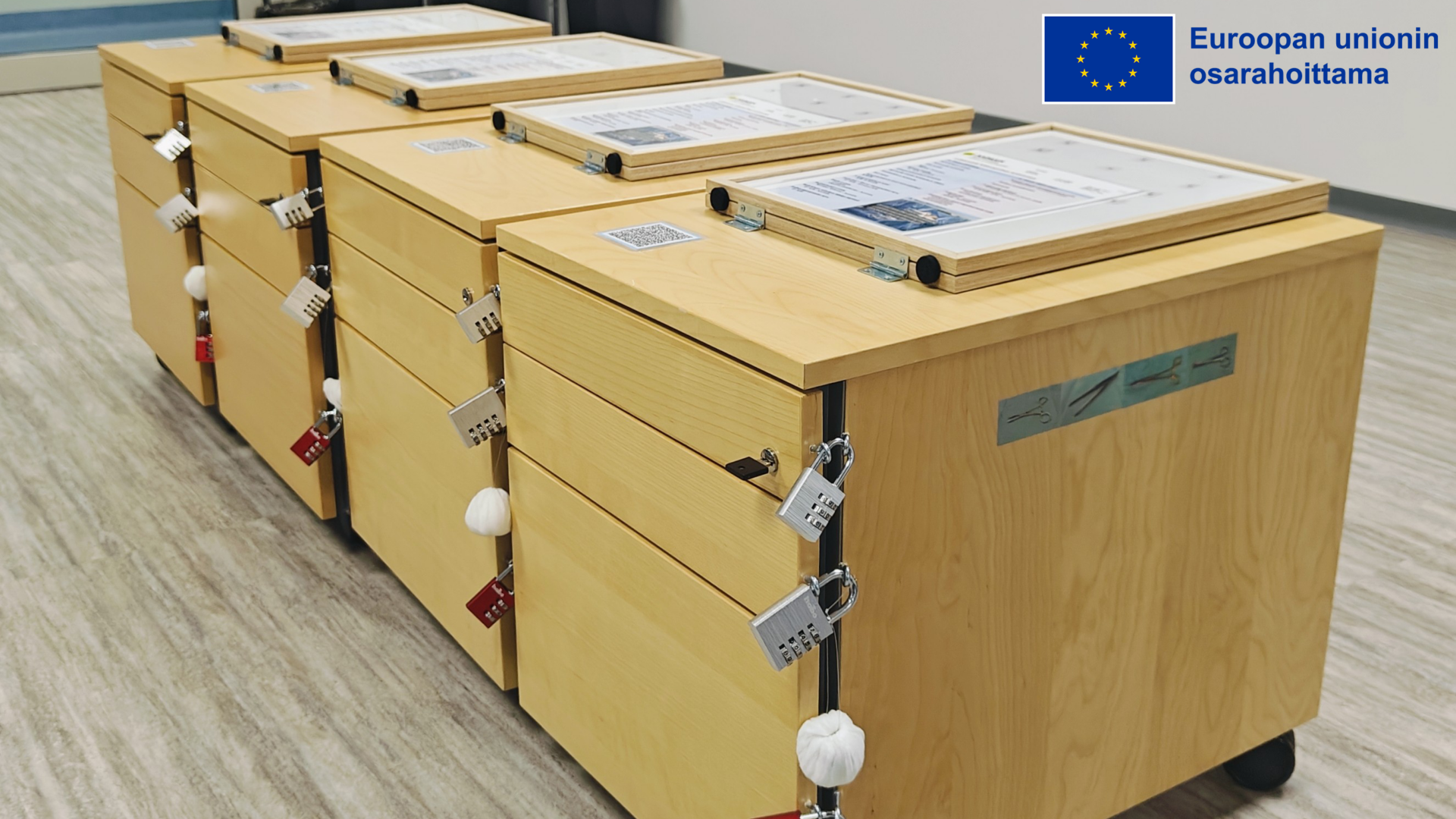 Neljä laatikostoa, jonka kaapit on lukittu numeroyhdistelmälukoilla. Kaappien päällä on paperiset ohjeet. Kuvan yläkulmassa Euroopan unionin osarahoittama -logo