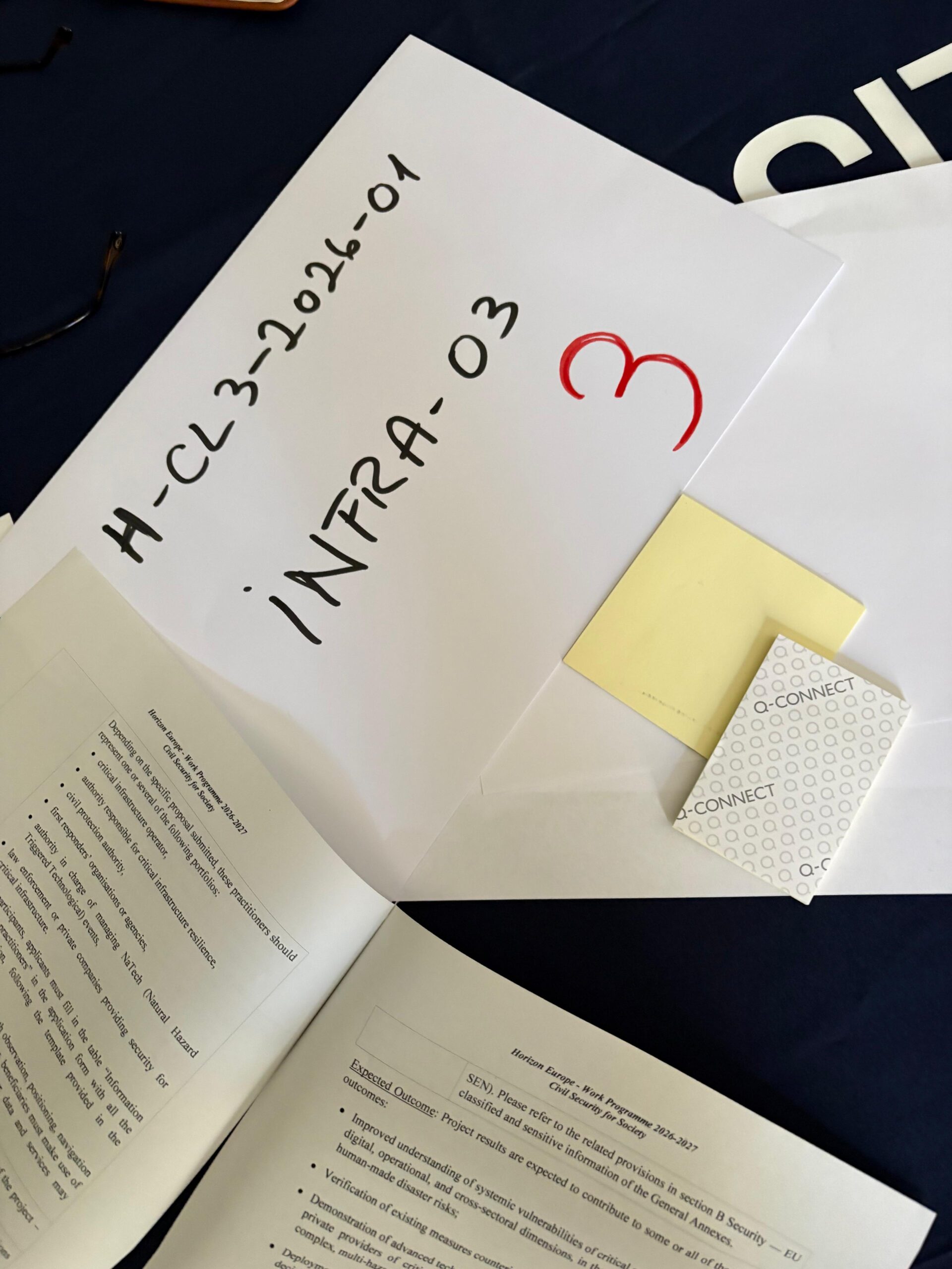 Multiple pieces of paper and notepads on a table. One paper has the words "H-CL3-2026-01 INFRA-03" written on it. Other papers have instructions written on them.