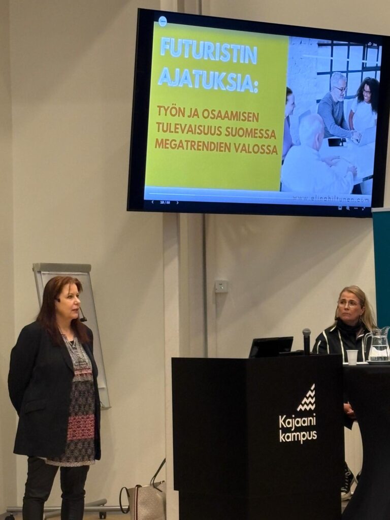 Kaksi naista esittelytilanteessa. Taustalla näyttö, jossa lukee "Futuristin ajatuksia: Työn ja osaamisen tulevaisuus Suomessa megatrendien valossa"