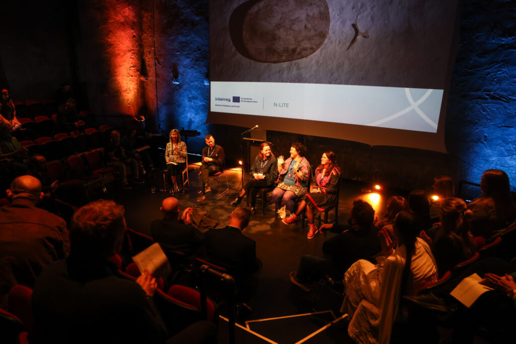 A photograph from a panel event. five people are on stage with people in the audience. In the background, a projector is displaying a slideshow with a Interreg Co-founded by the European union Northern Periphery and Arctic -logo and the N-LITE logo