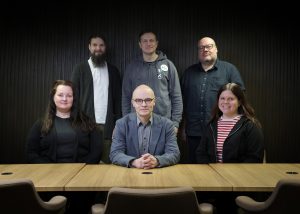 Kuusi henkilöä istuu ja seisoo pöydän takana hymyillen kameraan