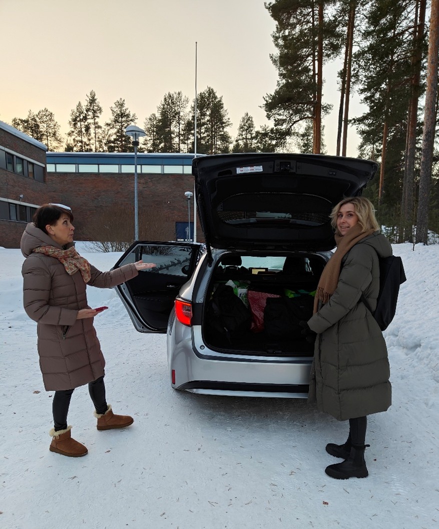 Kaksi naishenkilöä seisoo auton takana talvisessa pihassa. Auton ovet ja takaluukku ovat auki