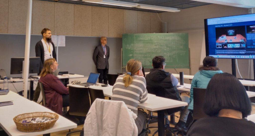 Kuva luokkatilasta, jossa kuuntelijat katsovat näyttöä jolla kuva videopelisivustolta. Kaksi henkilöä seisoo luokkatilan seinää vasten