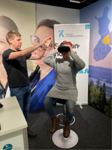Nainen istuu tuolill ja pitelee virtuaalilaseja päässään. Mies avustaa naista virtuaalilasien kanssa. Taustalla KAMKin rollup